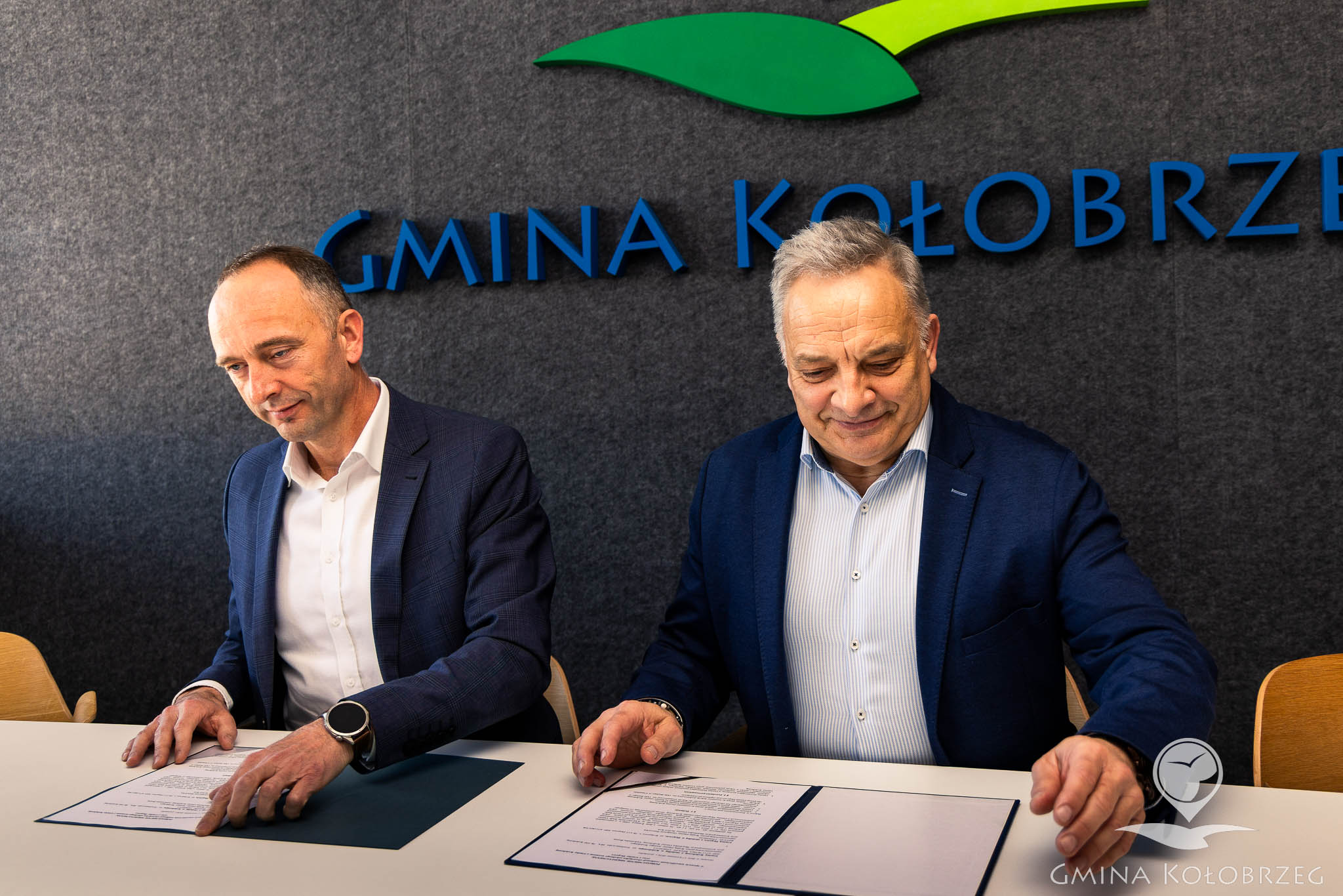 Dwaj mężczyźni siedzą przy stole i podpisują dokumenty; w tle napis &bdquo;Gmina Kołobrzeg&rdquo;. Na stole otwarte teczki z umową, scena przedstawia formalne podpisanie porozumienia lub dokument&oacute;w.