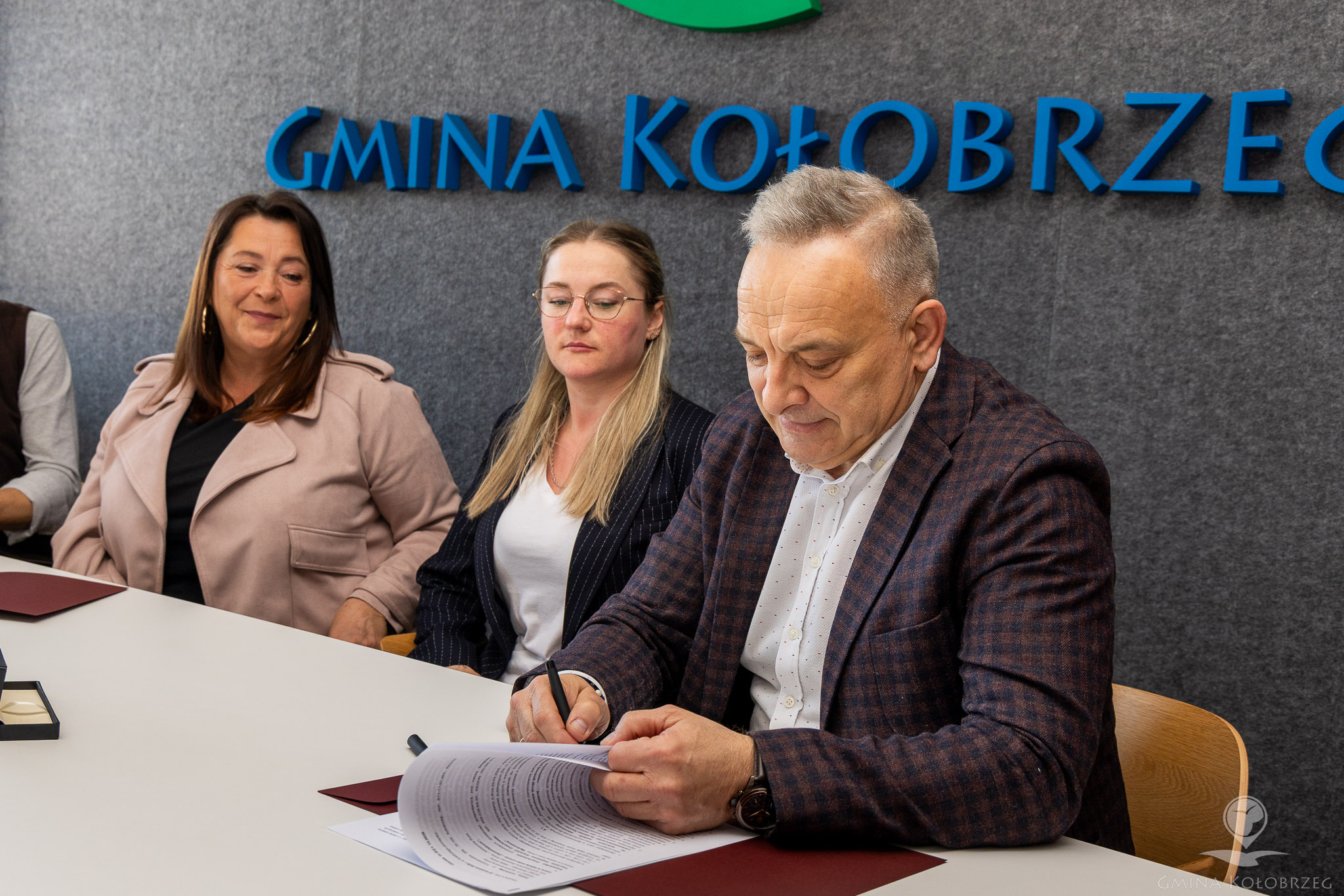 Zdjęcie z podpisania umowy Na zdjęciu widać mężczyznę podpisującego dokument przy stole, na tle napisu „Gmina Kołobrzeg”. Obok siedzą dwie kobiety obserwujące sytuację. Ujęcie ma charakter oficjalny i przedstawia moment zawierania umowy.