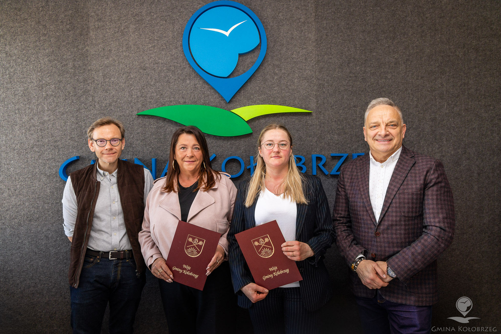 Zdjęcie z podpisania umowy Na zdjęciu widać cztery osoby stojące na tle ściany z logo „Gmina Kołobrzeg”. Dwie kobiety w środku trzymają dokumenty, a obok stoją dwaj mężczyźni. Ujęcie ma charakter oficjalny i dokumentuje ważne wydarzenie, podpisanie umowy.
