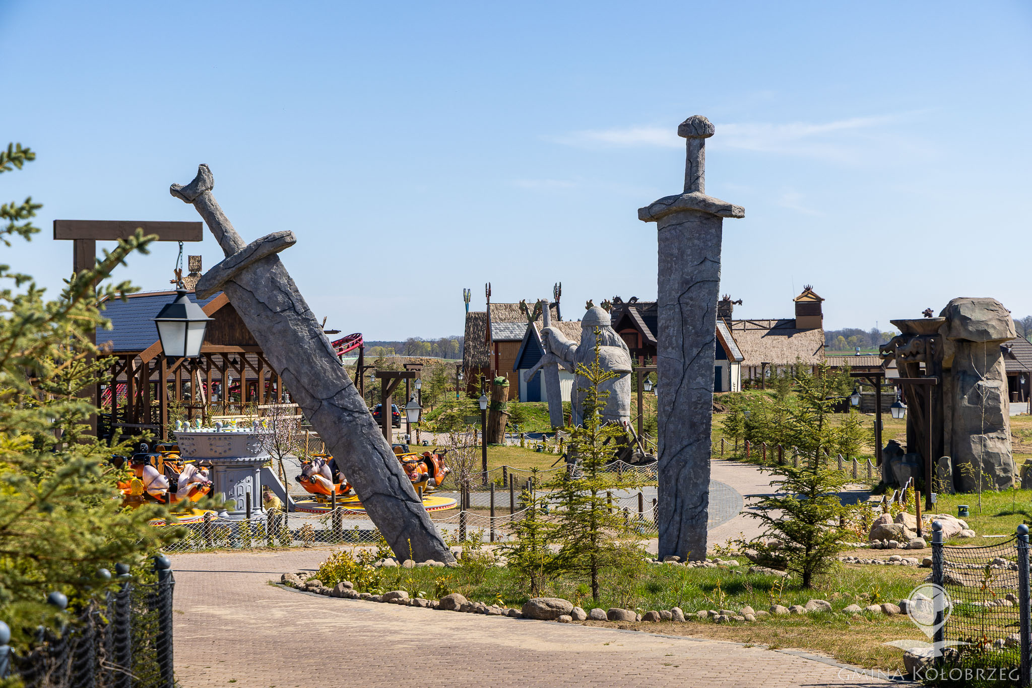 Hossoland Na zdjęciu widać teren parku rozrywki stylizowanego na klimaty historyczne lub fantastyczne. Na pierwszym planie znajdują się duże dekoracyjne miecze wbite w ziemię, a w tle widoczne są atrakcje, zabudowania tematyczne oraz karuzele. Całość otoczona jest zielenią i ścieżkami spacerowymi.