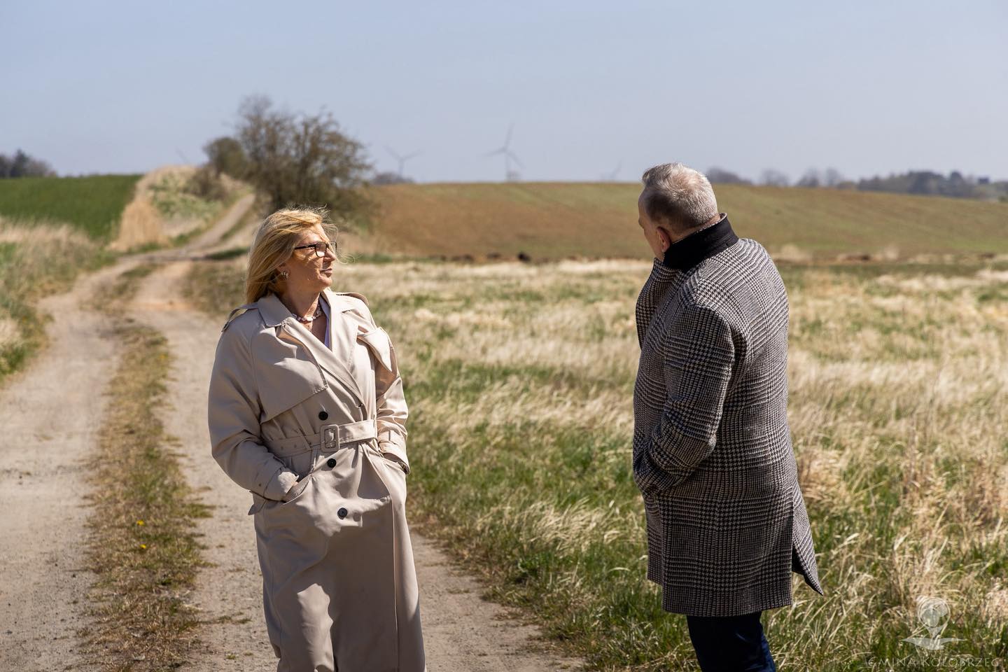 Zdjęcie z drogi Kobieta i mężczyzna stoją na polnej drodze i rozmawiają, w tle widać pola uprawne oraz turbiny wiatrowe na horyzoncie.
