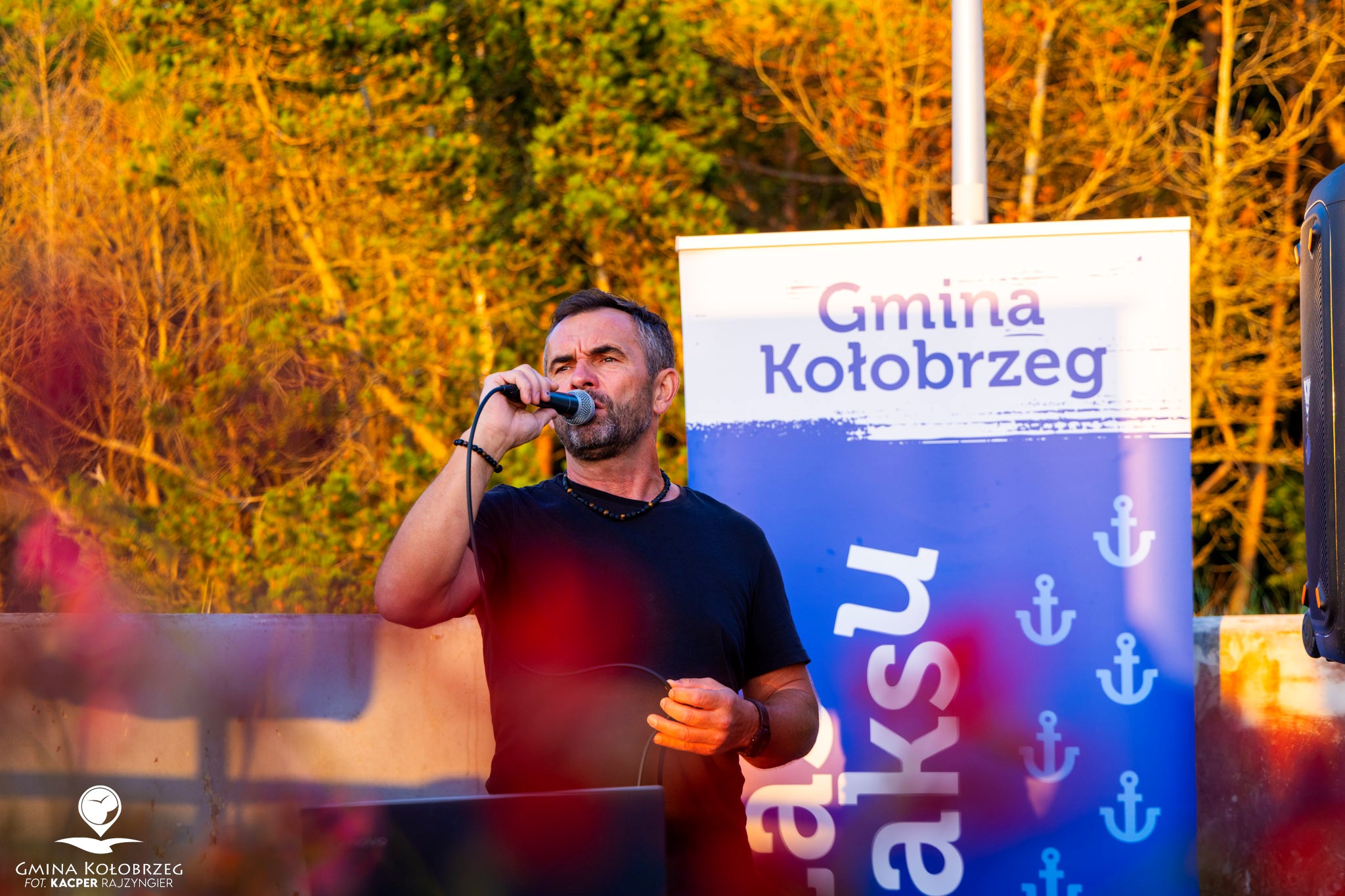 Zdjęcie przedstawia osobę prowadzącą wydarzenie plenerowe z mikrofonem. W tle widoczny jest baner &bdquo;Gmina Kołobrzeg&rdquo; z motywami wakacyjnymi oraz zieleń oświetlona ciepłym, wieczornym światłem.
