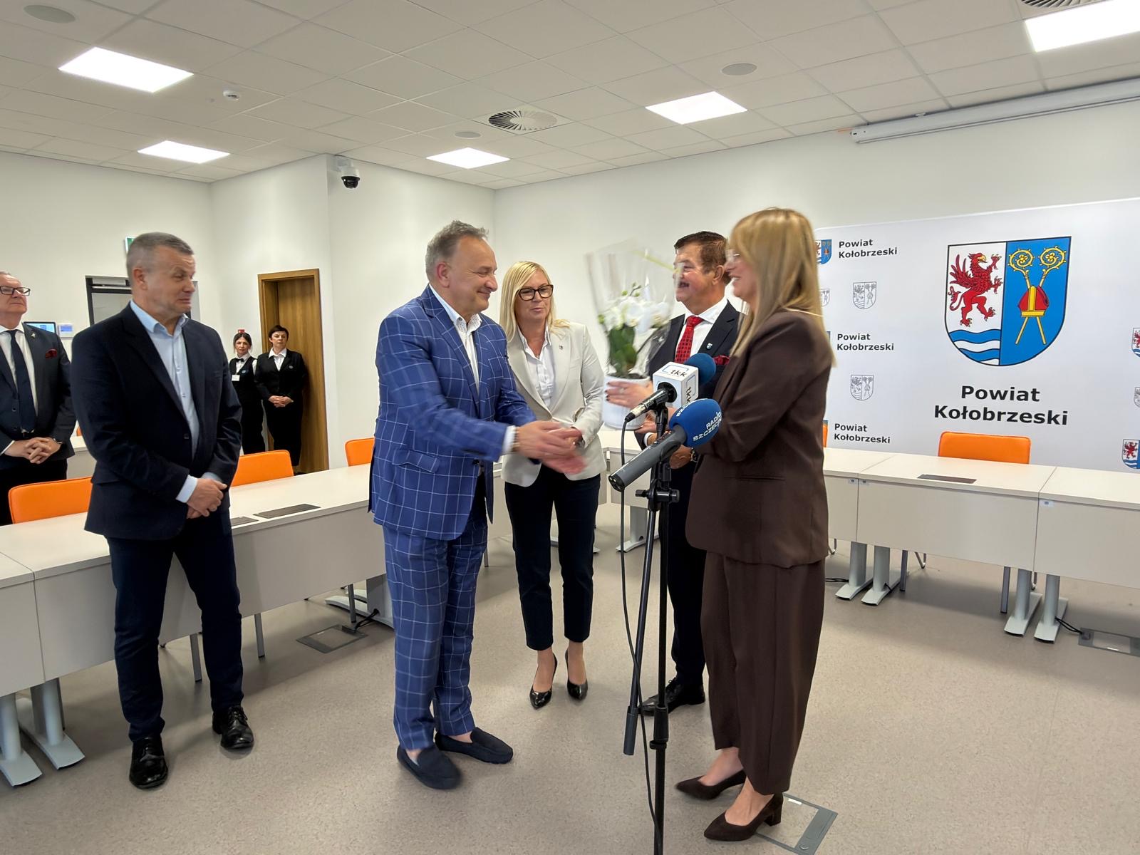 Zdjęcie z przekazaniem kwiatów Zdjęcie przedstawia oficjalne spotkanie w sali Powiatu Kołobrzeskiego. Widać przekazanie kwiatów przy mikrofonach, uczestników wydarzenia oraz ściankę z herbem i napisem „Powiat Kołobrzeski”.